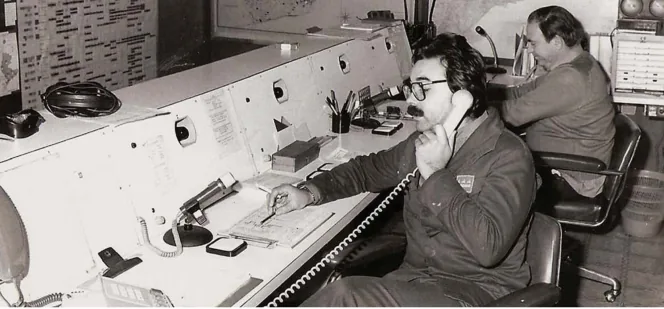 Fotografia en blanc i negre de dos operadors asseguts a la consola d'una sala de control: el del primer pla parla per un telèfon de fil mentre pren notes, al costat de micròfons de sobretaula i panells amb botons; al fons, un segon operador davant d'altres equips.
