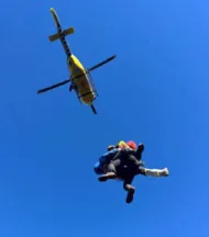 Helicòpter groc en vol, vist des de sota, amb un rescatador i una persona rescatada penjats de la grua de rescat.