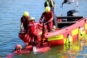 Bombers amb casc groc i armilla salvavides en una llanxa semirígida vermella i groga pugen a bord una persona que és a l'aigua, al costat d'un altre rescatador a l'aigua.