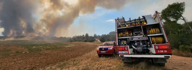Fotografia d'un camió de bombers vist per darrere (amb les persianes obertes, mànegues i material a la vista i el número 3.29.31) i d'un vehicle lleuger tot terreny aturats en un camí de terra entre camps, amb una gran columna de fum d'un incendi forestal al fons.