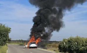 Fotografia d'un turisme blanc cremant a la vora d'una carretera en camp obert, amb flames a la part davantera i una gran columna de fum negre i dens.