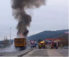 Fotografia d'un camió groc aturat en una via amb una gran columna de fum negre que surt de la part del darrere; a prop, vehicles de bombers i bombers treballant, i un senyal triangular d'avís a la calçada.