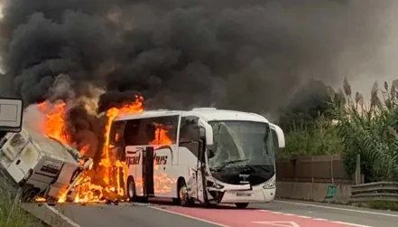Fotografia d'una carretera on un autocar blanc i una furgoneta bolcada al seu costat cremen amb flames intenses a la zona de contacte entre tots dos vehicles, amb una gran columna de fum negre.
