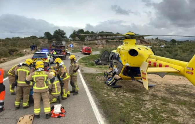 Fotografia en una carretera de muntanya: un grup de bombers amb uniforme i casc groc atén una víctima a terra al costat d'un helicòpter sanitari groc aterrat al marge de la via; al fons hi ha altres vehicles d'emergència amb els llums encesos.