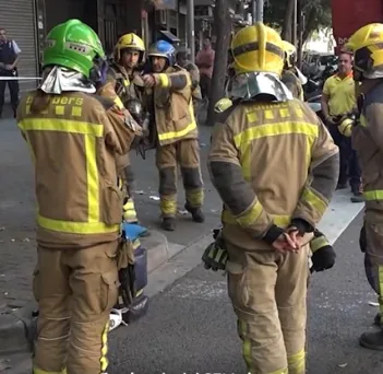 Fotografia en un carrer d'un grup de bombers d'esquena i de perfil, amb uniforme i cascos de diversos colors (verd, groc i blau), durant una intervenció urbana.