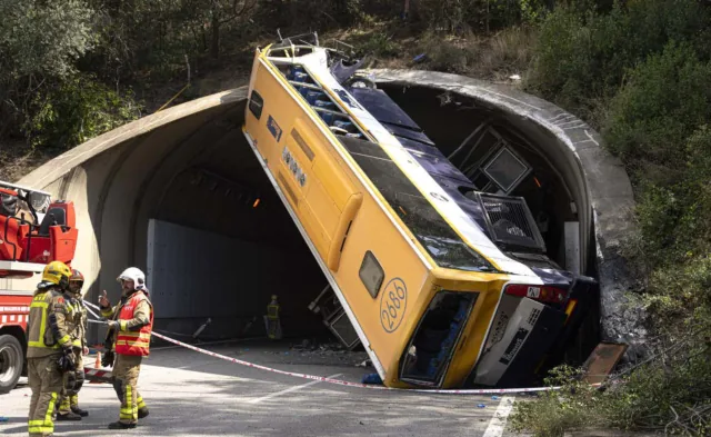 Fotografia d'un autobús groc bolcat i encastat en posició inclinada contra la boca d'un túnel, amb la part del davant enlaire i el sostre esventrat. En primer terme, bombers i un sanitari amb armilla taronja al costat d'un camió de bombers, darrere d'una cinta d'abalisament.