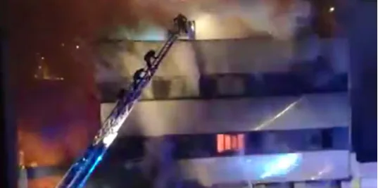 Fotografia nocturna d'un edifici industrial de diverses plantes en flames i ple de fum; una autoescala de bombers il·luminada s'estén fins a les plantes superiors, amb bombers enfilats al braç de l'escala.