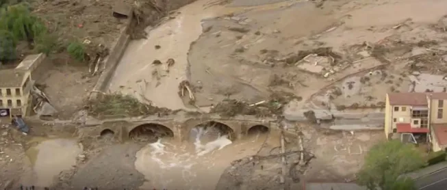 Fotografia aèria del riu Francolí després de la riuada: la llera i els marges coberts de fang, troncs i runa, un pont de pedra d'arcs amb l'aigua marronosa passant per sota, i edificis a banda i banda amb els baixos afectats.