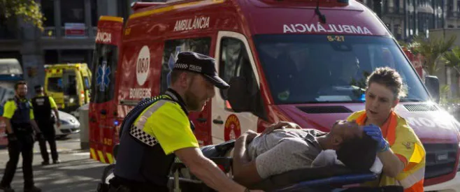 Fotografia d'un agent de la Guàrdia Urbana i una tècnica sanitària amb armilla que traslladen en llitera un ferit fins a una ambulància vermella (amb el rètol «Bombers» i el 060 al lateral); al fons, altres agents i vehicles d'emergència.