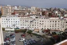 Fotografia de tres bombers amb jaqueta vermella i negra del GRAE (un d'ells amb el rètol «GRAE» a l'esquena) que caminen per la vorera carregant material, darrere d'una cinta d'abalisament policial; al fons, altres bombers i vehicles d'intervenció.
