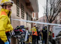 Fotografia del carrer tallat amb cinta policial: en primer terme un bomber amb roba groga; al fons, lliteres i material sanitari preparats, agents de policia i personal amb armilla reflectant al costat de l'edifici afectat, i una ambulància aparcada.