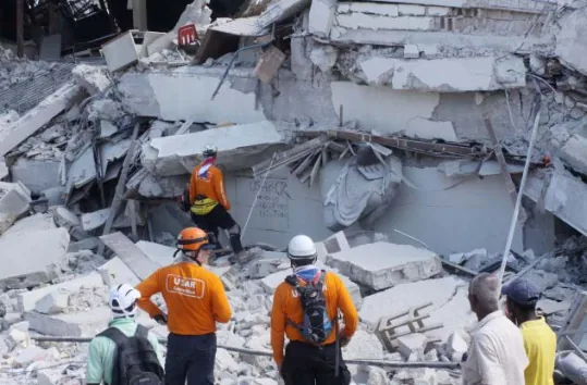 Fotografia d'un edifici de formigó completament ensorrat, amb lloses trencades i runa amuntegada. Diversos rescatadors amb casc i samarreta taronja (un d'ells amb el rètol «USAR» a l'esquena) inspeccionen la runa, acompanyats d'altres persones; un rescatador s'enfila pels blocs de formigó al centre de la imatge.