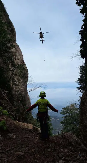 Fotografia vertical presa des d'un coll estret entre dues parets de roca: un bomber del GRAE, d'esquena, amb casc i armilla d'alta visibilitat, fa senyals amb els braços estesos a un helicòpter que vola en estacionari al damunt, amb el cable del gruatge penjant.