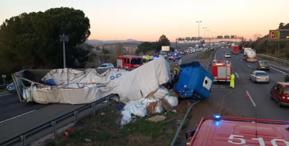 Fotografia al capvespre d'una autovia amb un camió sinistrat al marge: la cabina blava ha quedat separada i tombada i la caixa, amb la lona estripada, ha bolcat i ha escampat sacs de càrrega per la cuneta. Al voltant hi ha diversos vehicles de Bombers amb els llums d'emergència encesos i personal treballant, mentre el trànsit continua circulant pels carrils del costat.